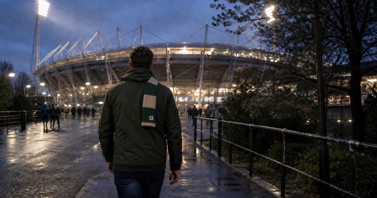A football supporter wearing a green scarf walking towards a floodlit stadium